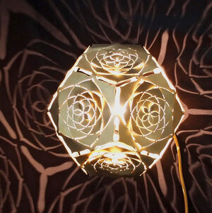 Platonic Solid: "The Sun" and its Light Patterns An image of 'The Sun' lamp illuminated in a dark room, showcasing its intricate brass design and warm radiance. Inspired by the beauty of the dodecahedron and the intricate patterns found in flowers, each of the sculpture's twelve faces features rounded cuttings that resemble the petals of a flower, creating an optical illusion that is nothing short of mesmerizing. Crafted from 1.5mm thick brass material.